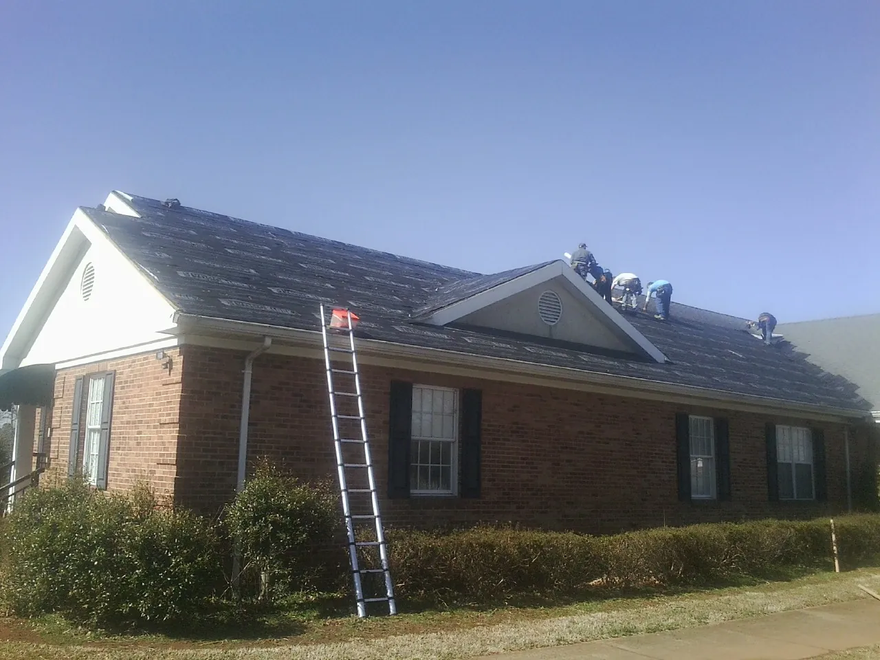 Crew tearing shingles off a brick ranch roof with a ladder set — Emergency Roof Repair work by our crew in Mesick