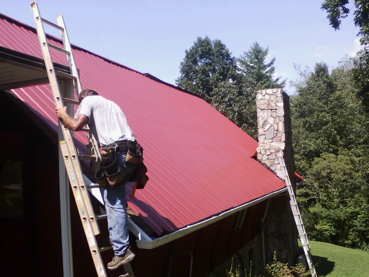 Roofer on a ladder beside a red metal roof and stone chimney — Soffit & Fascia Repair by Roofing Brothers in Mesick